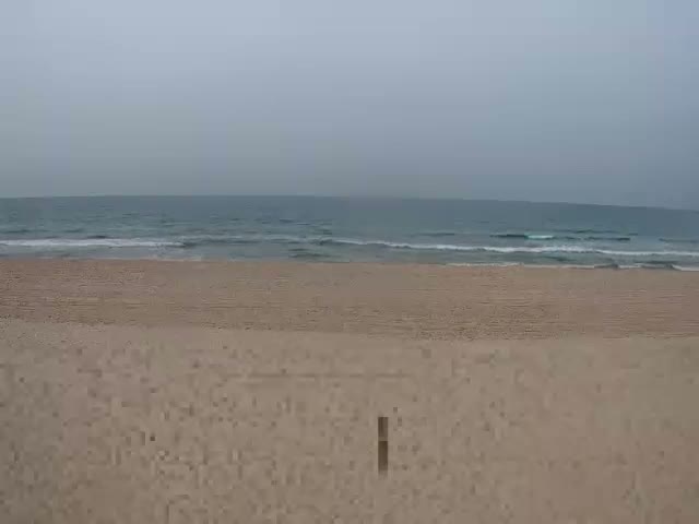A wide, light brown sandy beach meets a blue-green ocean with gentle waves under a cloudy, overcast grey sky.