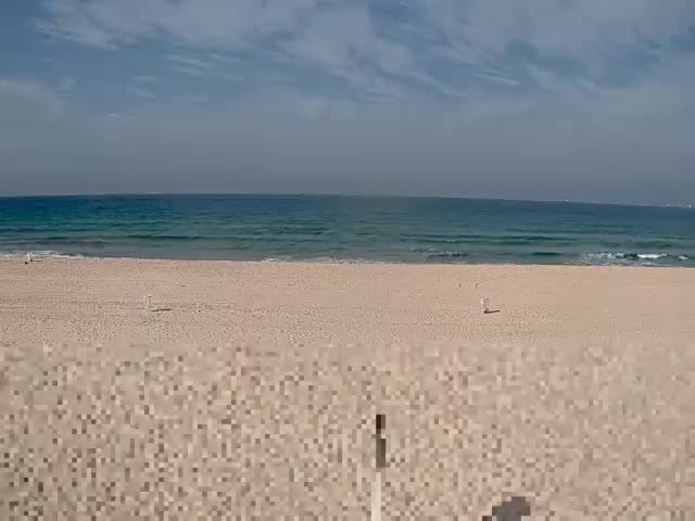 A sandy beach stretches towards a blue ocean with gentle waves under a partially cloudy sky, indicating fair weather.