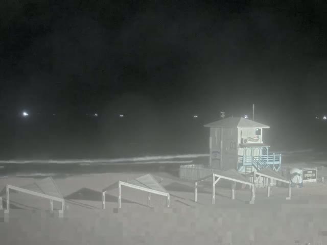 A lifeguard stand sits on a hazy, dark beach at night, with several small structures and a calm ocean visible in the distance.