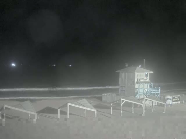 A lifeguard stand sits on a hazy, dark beach at night, with several small structures and a calm ocean visible in the distance.
