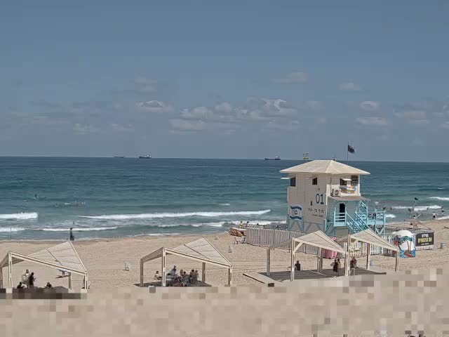 A sandy beach with people relaxing under shade structures, a lifeguard tower, and small waves rolling onto the shore under a partly cloudy sky.