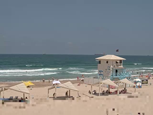 A sandy beach with a lifeguard stand, several beach umbrellas, and people enjoying a sunny day with moderate waves rolling in on the ocean.