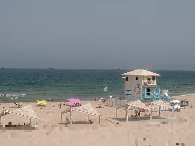 A sandy beach with light-colored structures, several people, and a lifeguard station is visible on a partly cloudy day, with calm waves lapping the shore and a few ships in the distance.