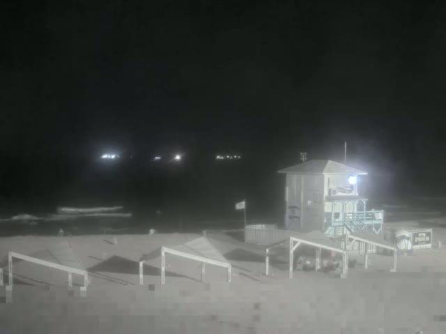 A nighttime beach scene shows a lifeguard stand and several beach shelters under a dark sky with a calm ocean in the background.