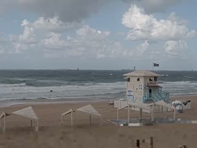 A lifeguard tower stands on a sandy beach with several canopies, facing a choppy ocean dotted with distant ships under a partly cloudy and windy sky.