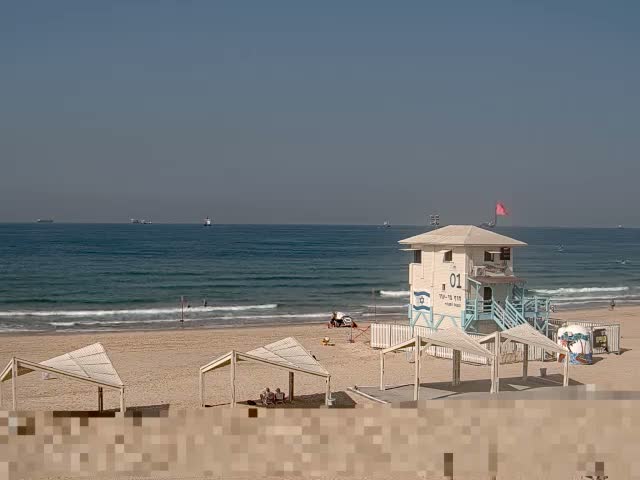 A lifeguard tower stands on a sandy beach with several canopies, facing a choppy ocean dotted with distant ships under a partly cloudy and windy sky.