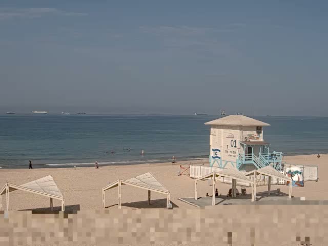 A sunny beach features a prominent white and blue lifeguard tower and several open-sided shelters, with a few people on the sand and in the calm blue sea, where distant ships are visible under a bright, clear sky.