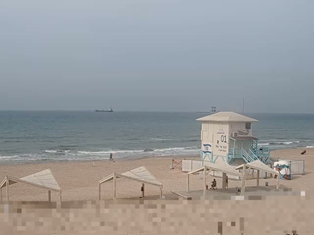 A wide view of a sandy beach features a white lifeguard tower with light blue stairs, several open shelters, a few people, and a distant cargo ship on the calm sea under an overcast sky.