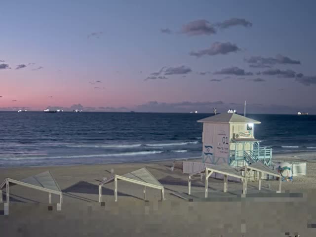 A tranquil beach scene at twilight features a lit lifeguard tower and several open shelters on the sand, with gentle waves washing ashore and distant ships visible on the horizon beneath a partly cloudy sky transitioning from blue to pink.