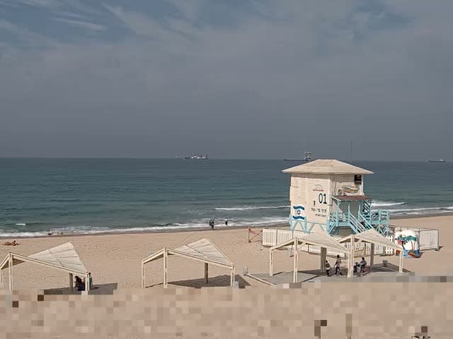 A sandy beach features a white and light blue lifeguard tower, several shaded lounge areas, and a few people enjoying the calm, blue-green sea under an overcast sky, with distant ships visible on the horizon.