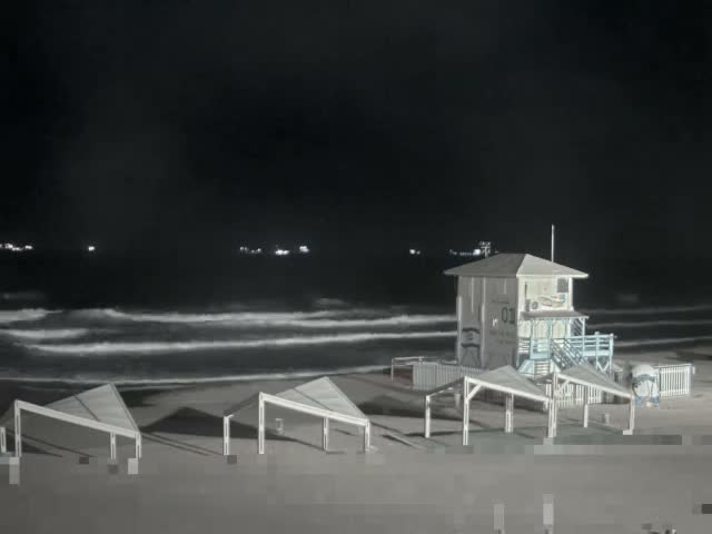 A lifeguard tower and several white-framed beach shelters stand on the sand at night, with waves breaking on the shore and distant lights visible on the dark ocean under a clear sky.