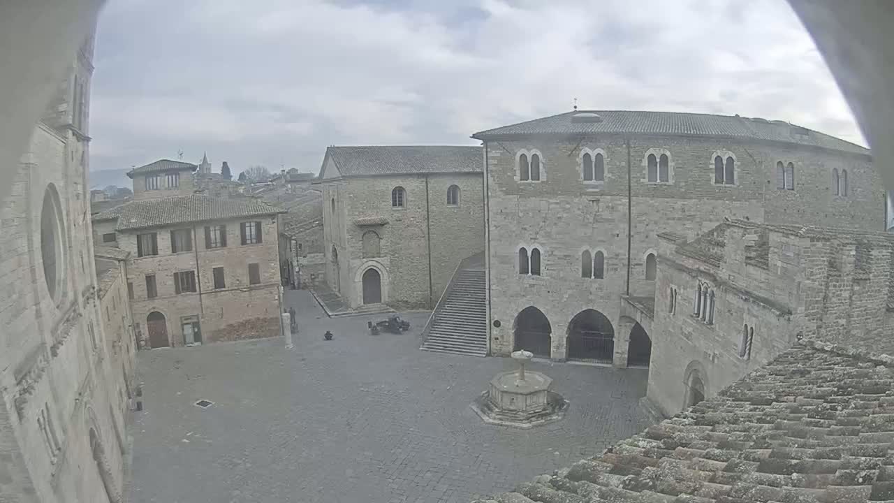 A view from a rooftop shows an overcast day in a stone plaza surrounded by medieval-style buildings, one featuring a fountain in its center.