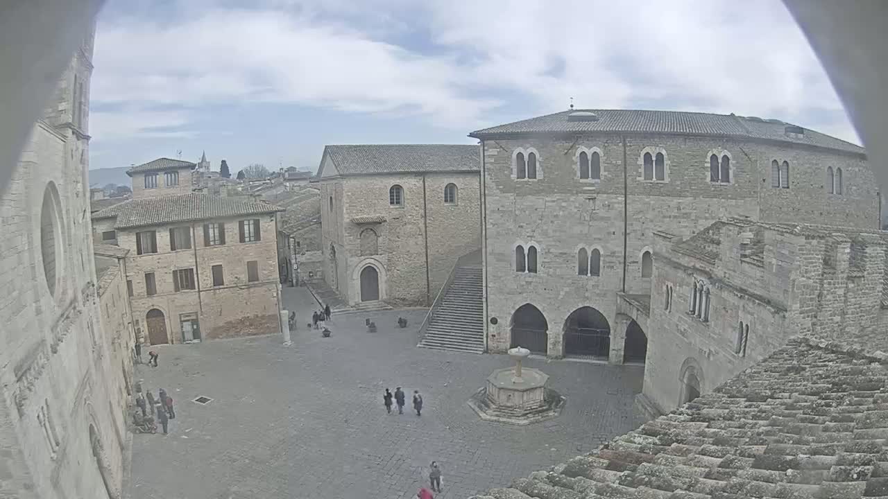 A high-angle view of a town square with several stone buildings, a central fountain, and a few people, under a cloudy sky.