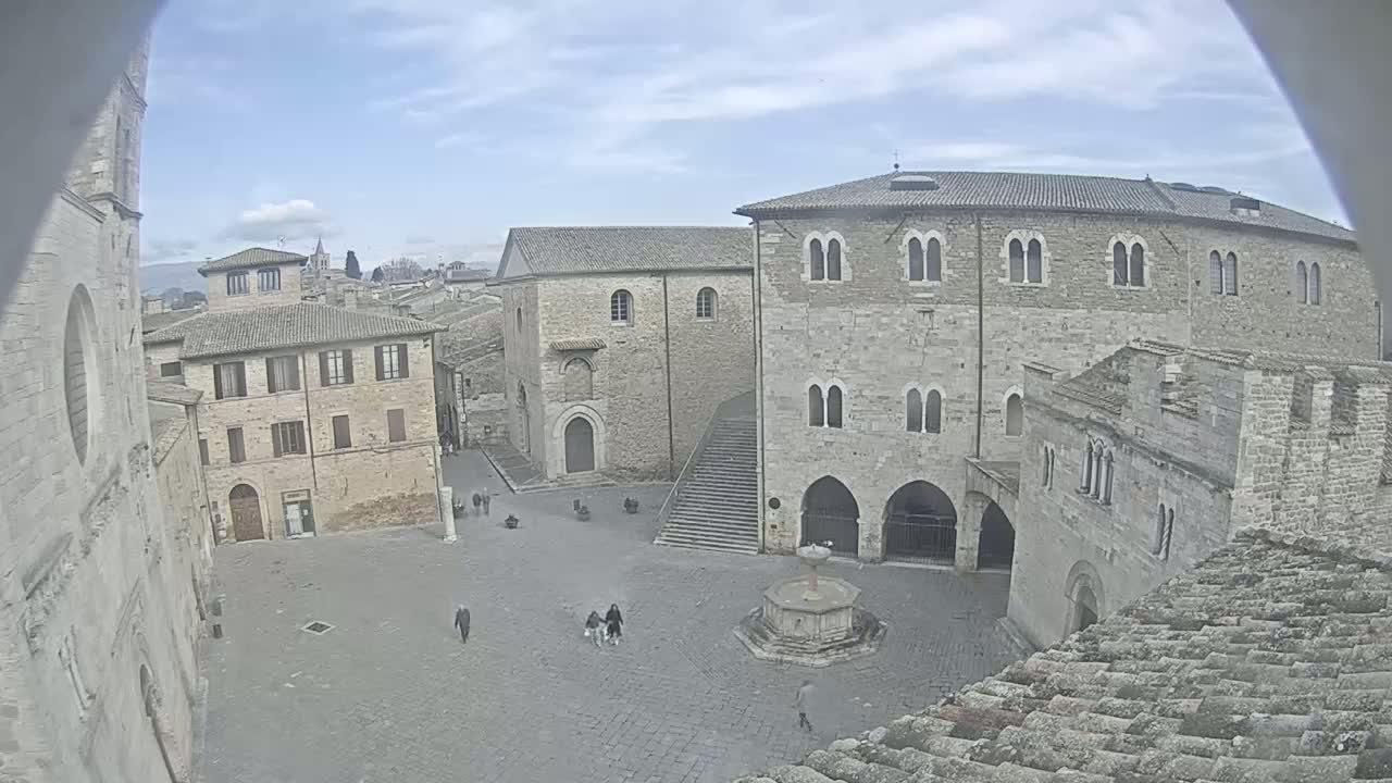 A mostly empty stone plaza, surrounded by medieval-style buildings, under a partly cloudy sky.