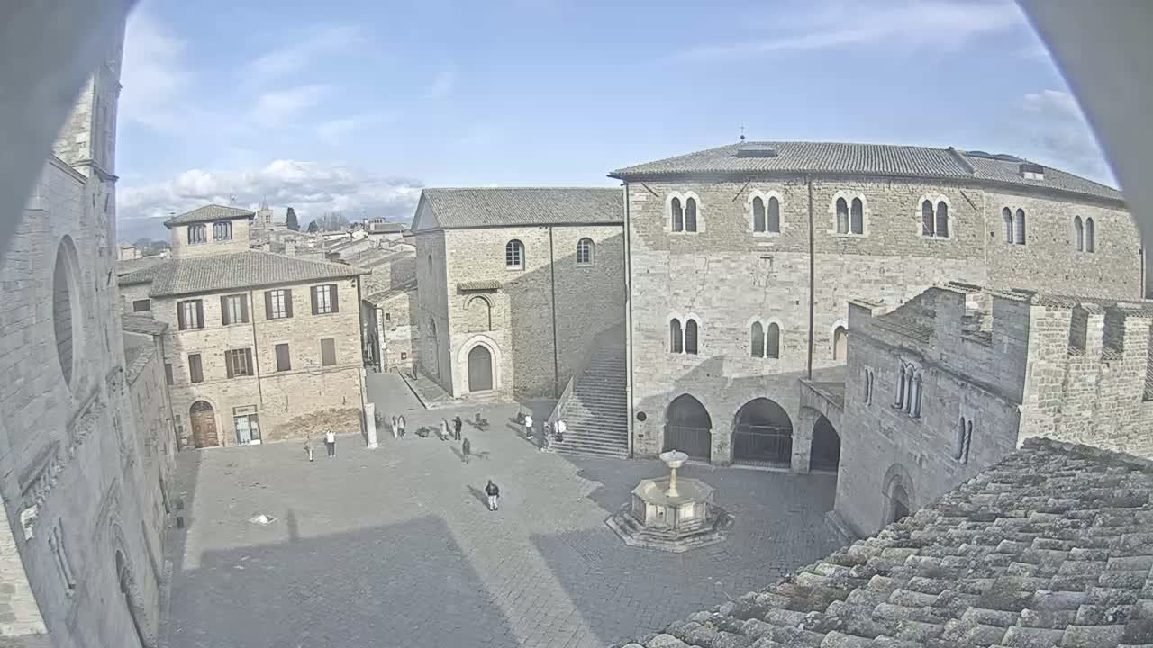 A mostly sunny day shows a cobblestone town square surrounded by several stone buildings, one with a fountain at its center, and a few people walking around.