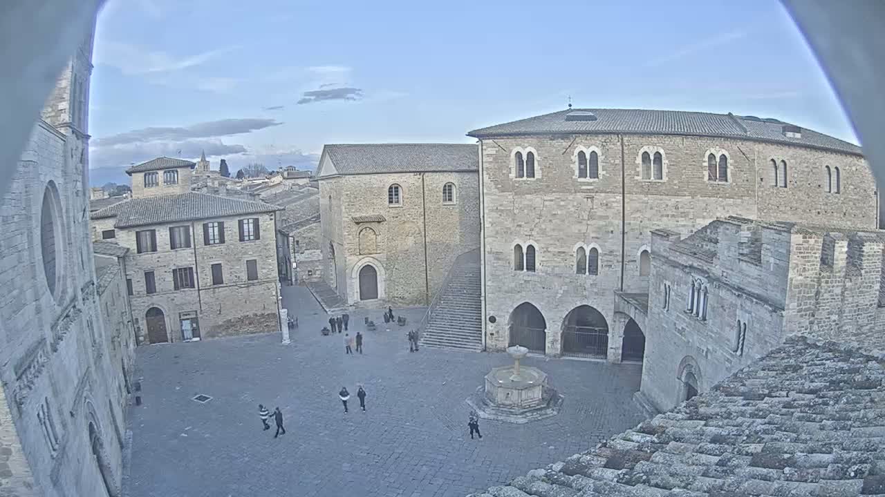A mostly empty town square with several stone buildings, a fountain, and a few people walking around on a partly cloudy day.