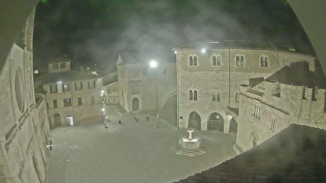 A nighttime view of a town square with stone buildings, a fountain, and a hazy atmosphere.