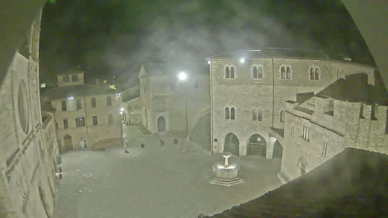 A hazy night scene shows a town square with several stone buildings surrounding a fountain.