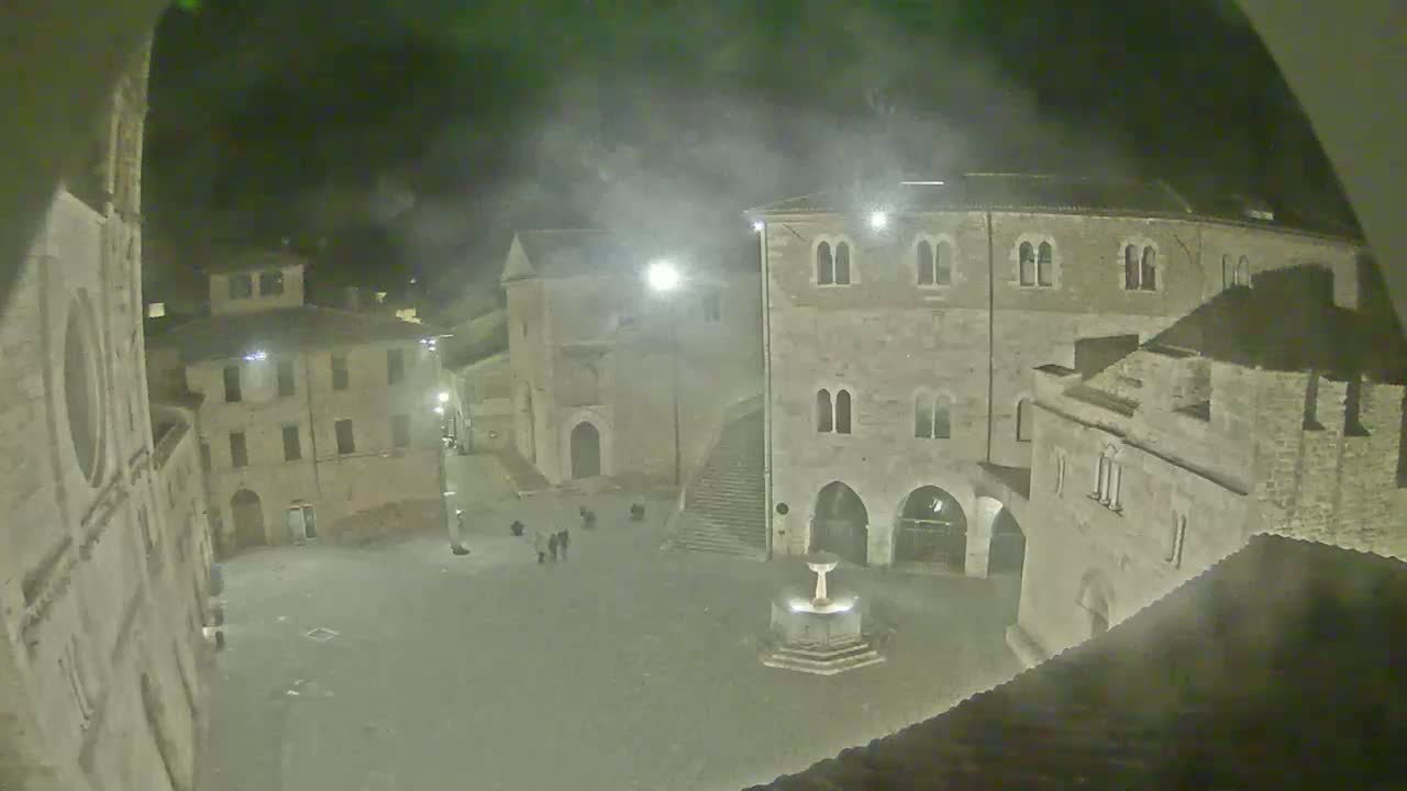 A nighttime view of a town square with several stone buildings surrounding a fountain, under hazy or foggy conditions.