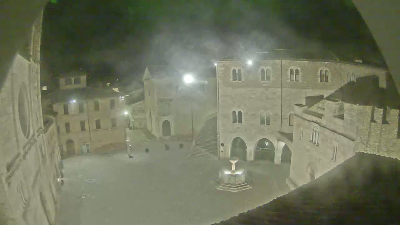A nighttime view of a town square with stone buildings surrounding a fountain, under hazy conditions.