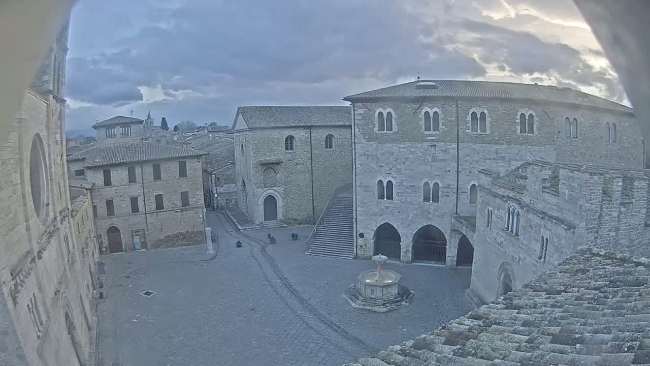 A mostly empty town square with a fountain, surrounded by old stone buildings under a cloudy sky.