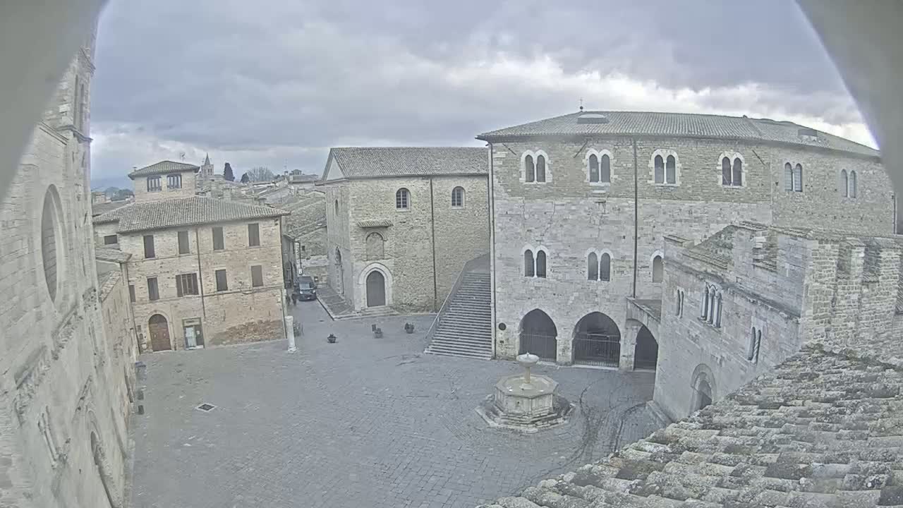 A stone plaza with a fountain is surrounded by medieval-style buildings under a cloudy sky.
