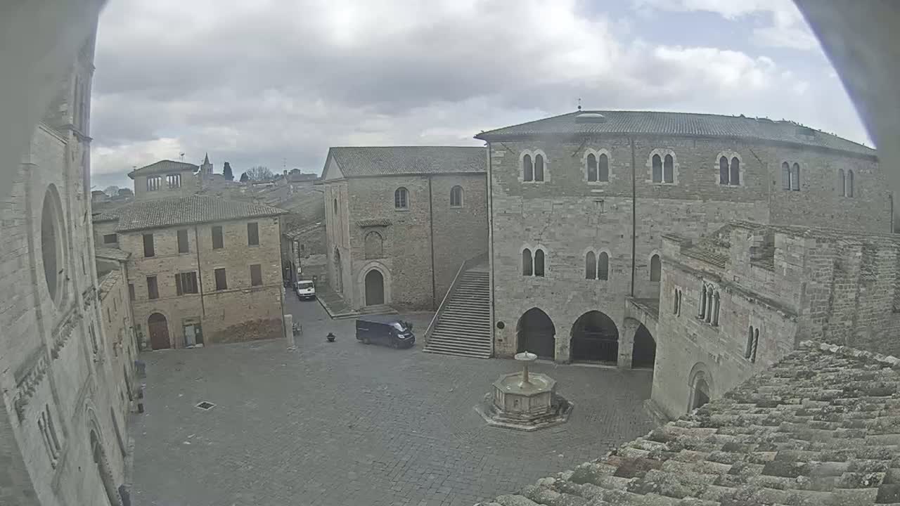 A large, stone plaza is seen on an overcast day, surrounded by medieval-style buildings, including a fountain in the center, and a single vehicle parked to the side.