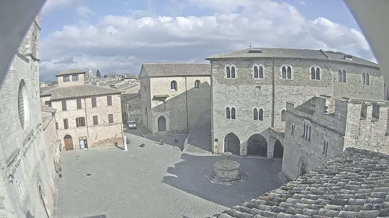 A partly cloudy day reveals a town square surrounded by medieval-style stone buildings, a fountain at its center, and a view of additional buildings in the distance.