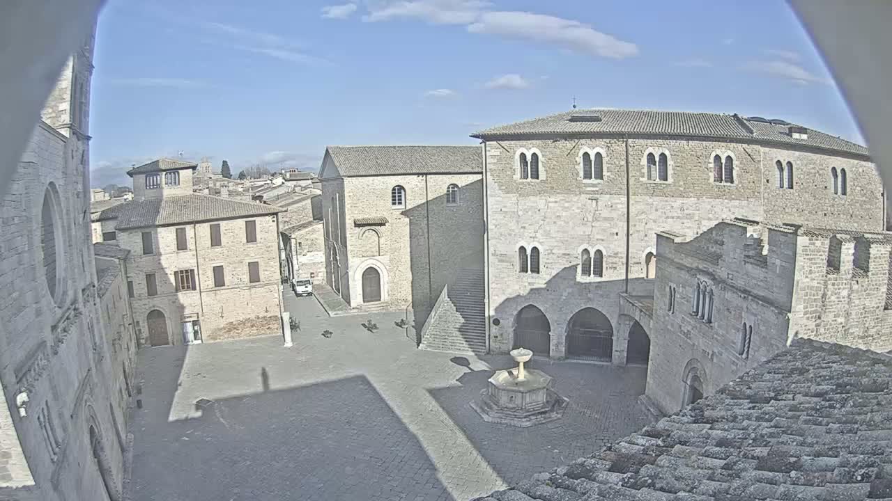 A sunny day reveals a stone courtyard in a historic town, featuring several aged buildings surrounding a central fountain.