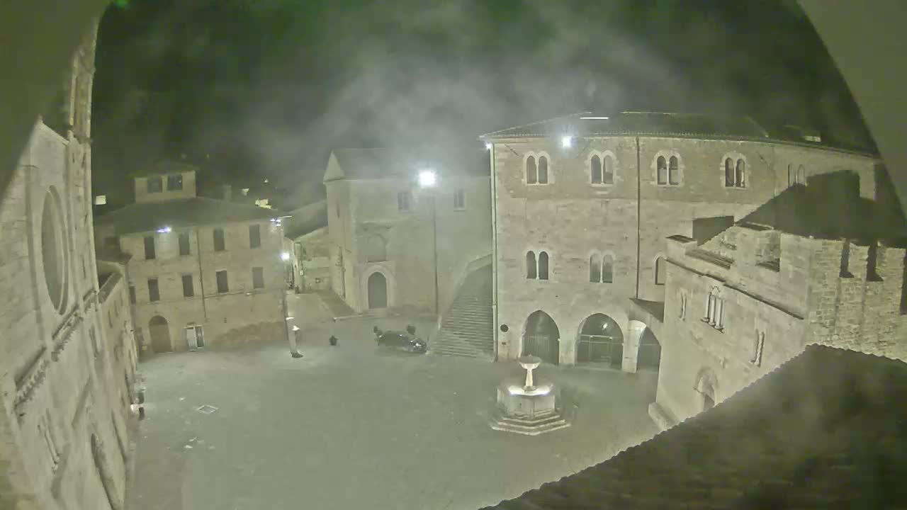 A fog-shrouded town square at night, featuring several stone buildings surrounding a central fountain.