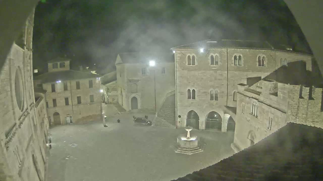 A nighttime view of a town square with several stone buildings surrounding a fountain, under foggy conditions.