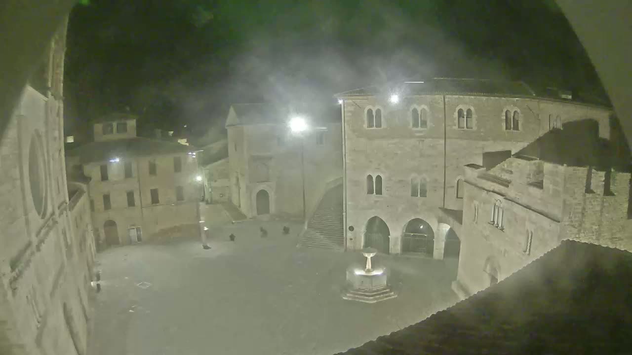A nighttime, hazy view of a town square with several stone buildings surrounding a fountain.