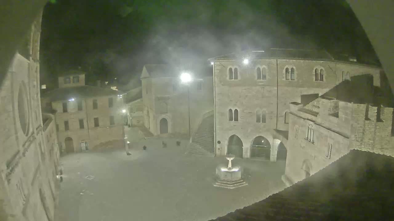 A nighttime view of a town square with stone buildings surrounding a central fountain, under hazy or foggy conditions.