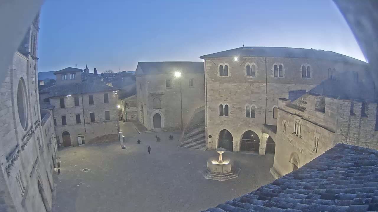 A mostly empty town square at dusk, surrounded by stone buildings, features a fountain in the center.