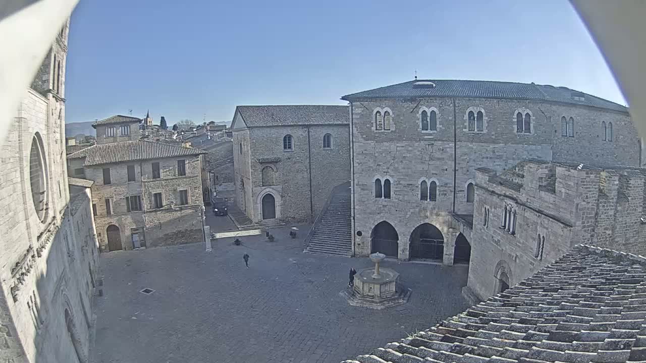 A sunny day reveals a town square surrounded by stone buildings, including a large structure with arched entryways and a central fountain.