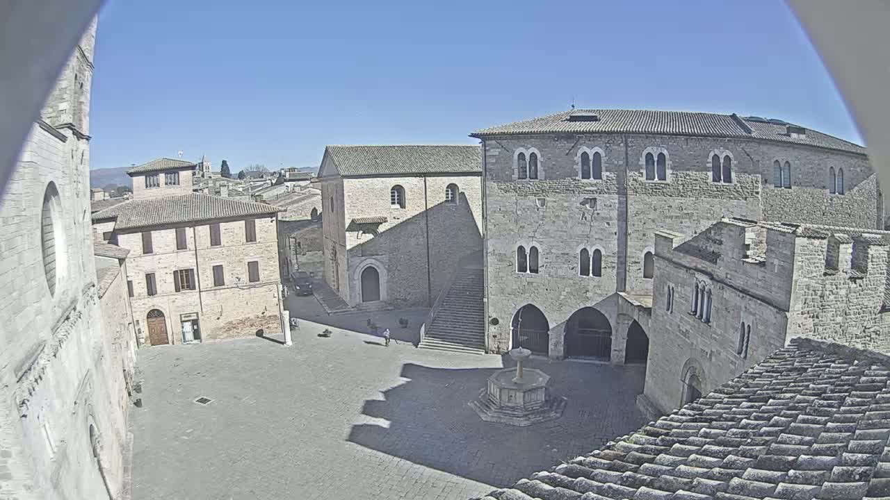 A sunlit town square, surrounded by stone buildings, features a central fountain and a few pedestrians under a clear blue sky.