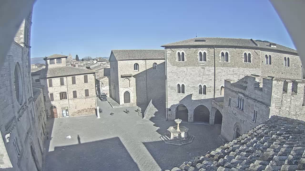 A stone plaza in a medieval-looking town on a sunny day, centered around a fountain, with several buildings surrounding the open space.