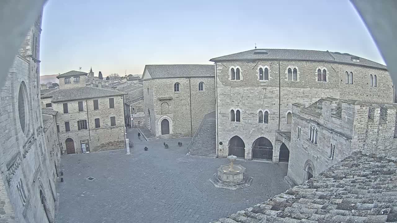 A mostly empty stone plaza is surrounded by ancient-looking buildings under a clear, light-blue sky.