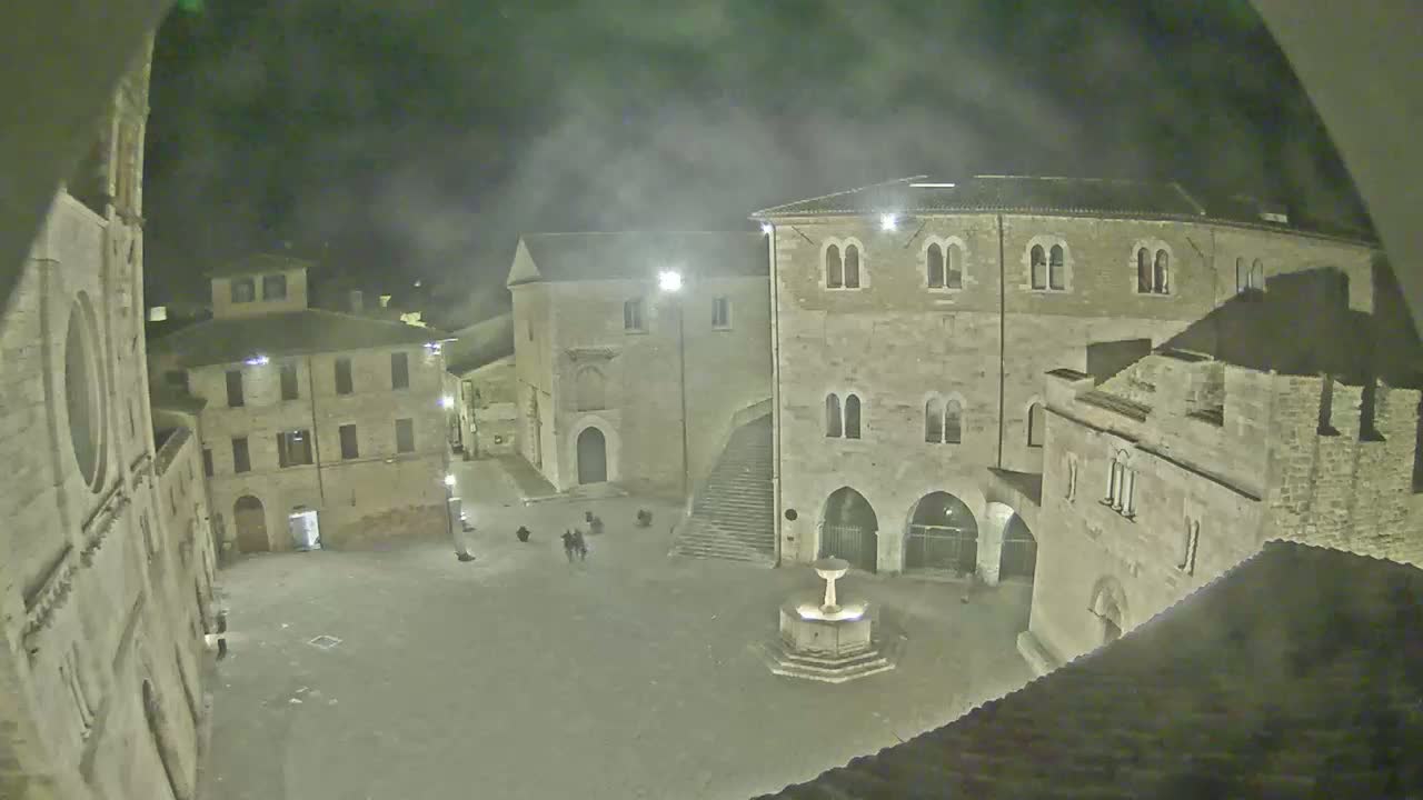 A nighttime view of a town square, under a hazy sky, shows several stone buildings surrounding a central fountain with a few people in the square.