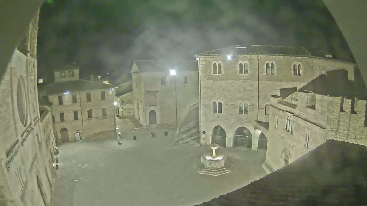 A nighttime view of a stone plaza in a historic town, under hazy or foggy conditions, with a fountain in the center and several buildings surrounding it.