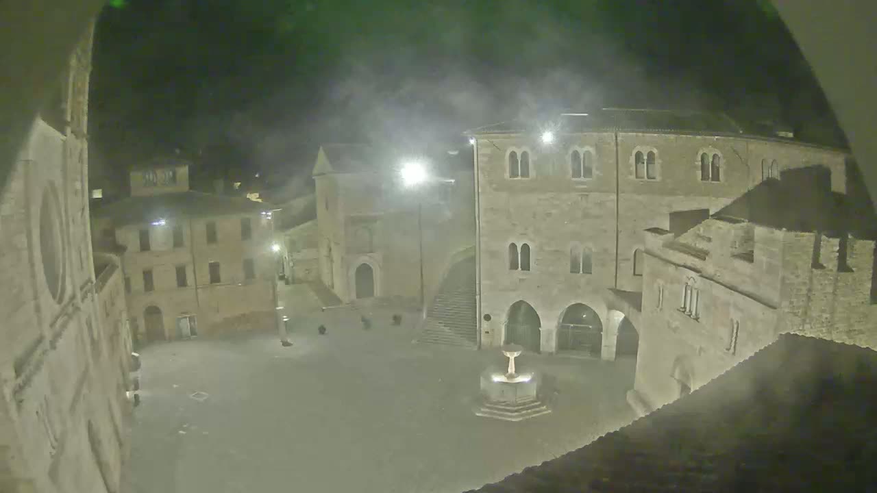 A nighttime view of a town square with stone buildings and a fountain, under hazy or foggy conditions.