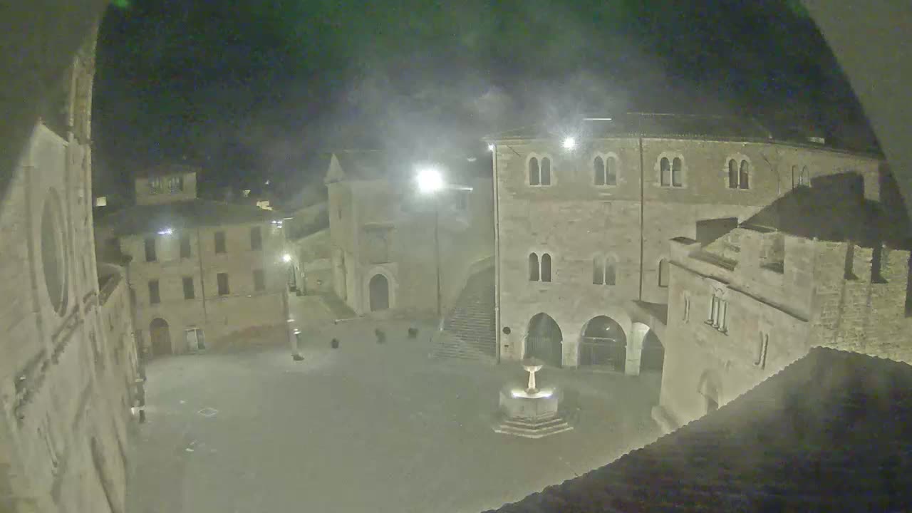 A nighttime view of a town square with stone buildings surrounding a central fountain, under hazy or foggy conditions.