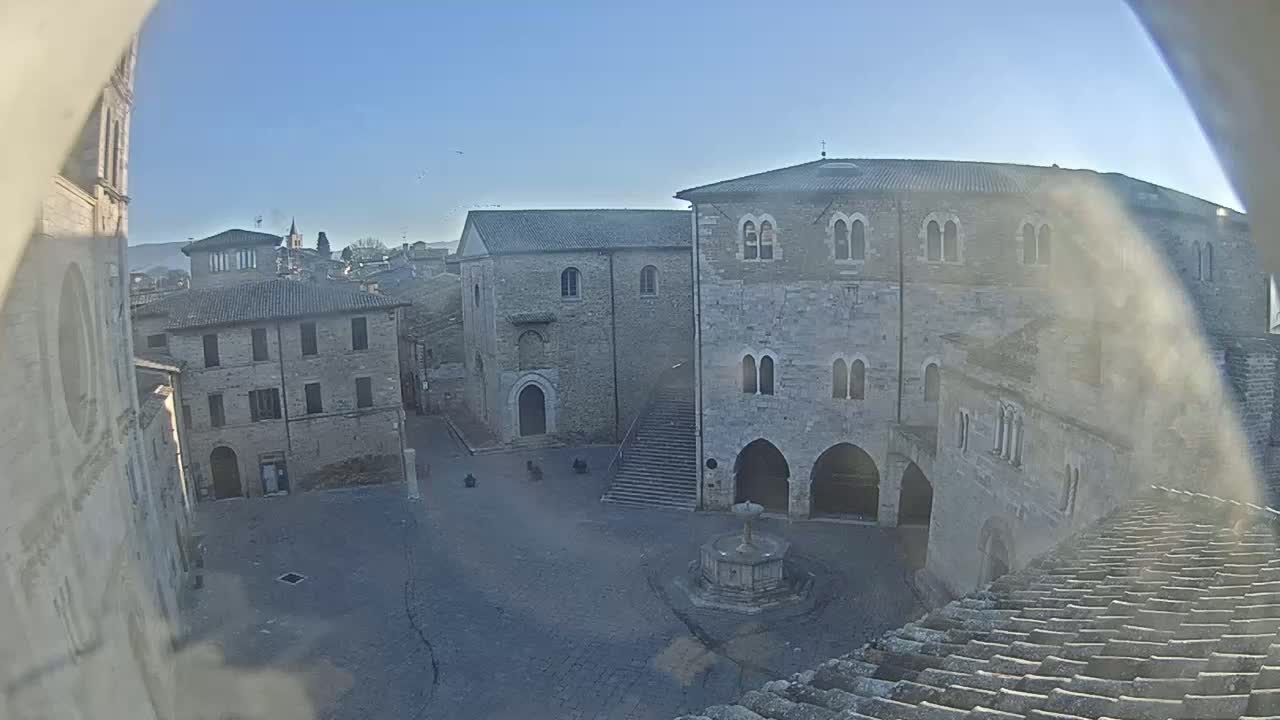 A mostly empty town square with a fountain, surrounded by aged stone buildings under a clear blue sky.