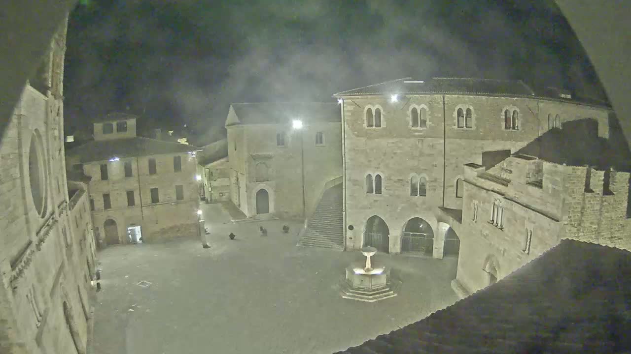 A nighttime view of a town square with stone buildings surrounding a central fountain, under a hazy, dark sky.