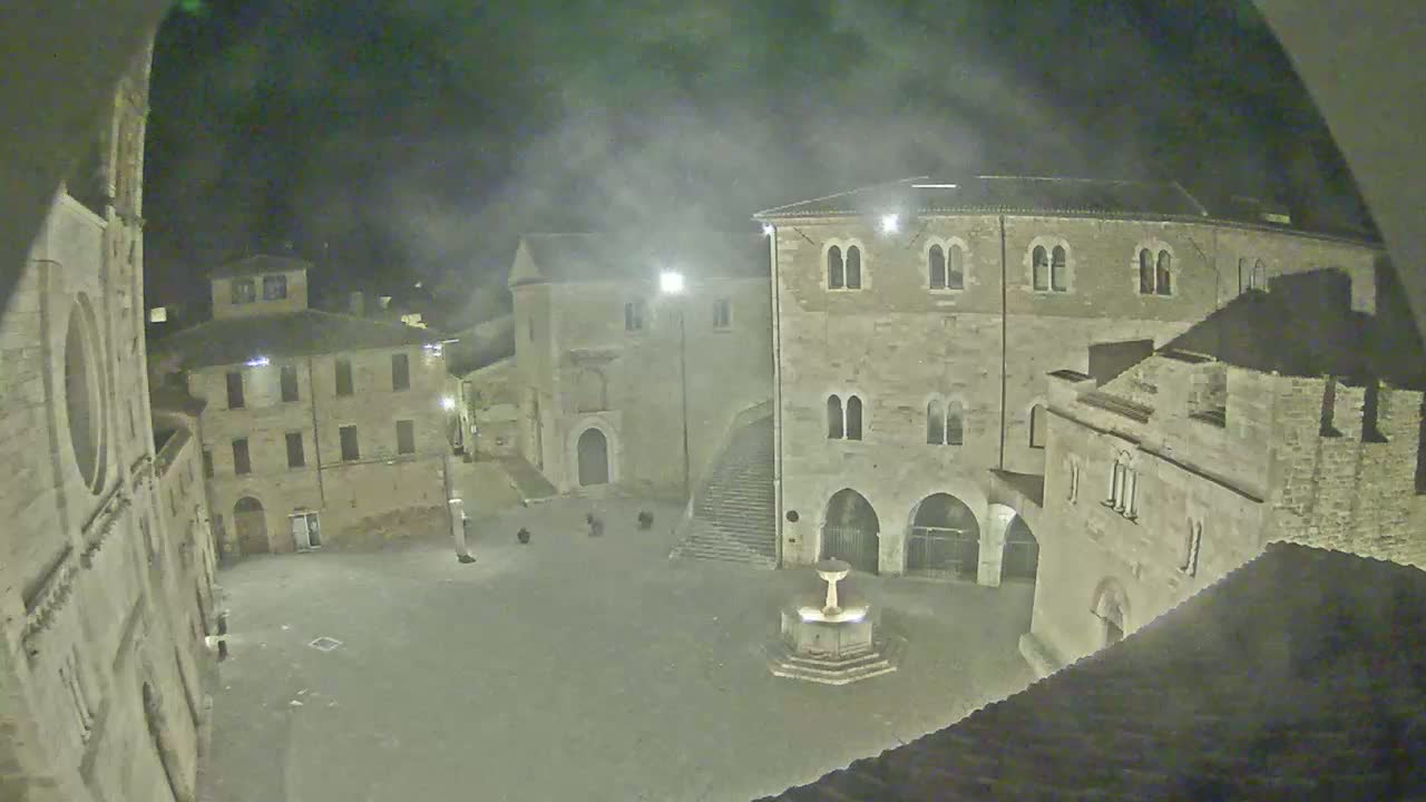 A nighttime view of a town square with several stone buildings surrounding a central fountain, under a hazy or foggy sky.