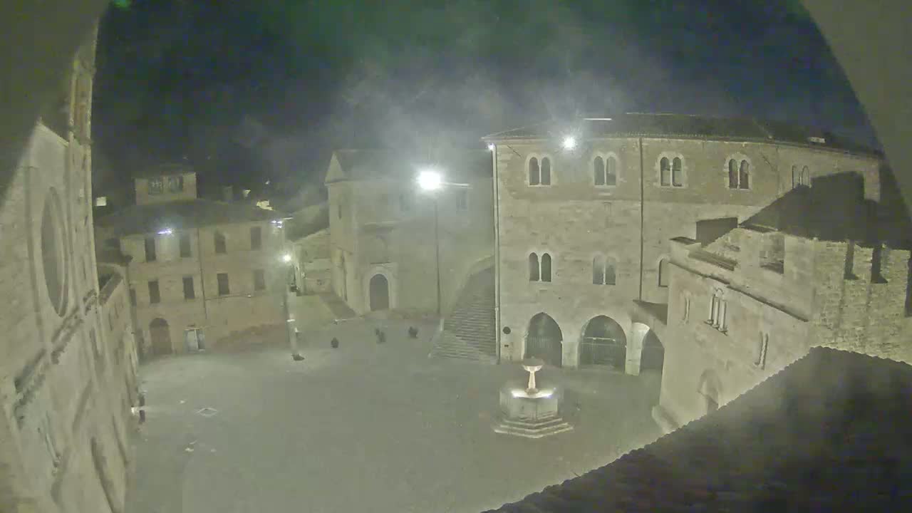 A nighttime view of a town square with several stone buildings surrounding a central fountain, under hazy conditions.