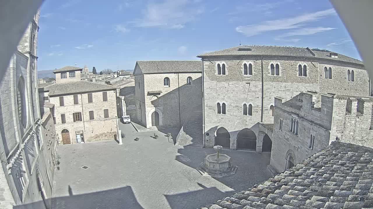 A stone plaza with a fountain is surrounded by medieval-style buildings under a sunny sky.
