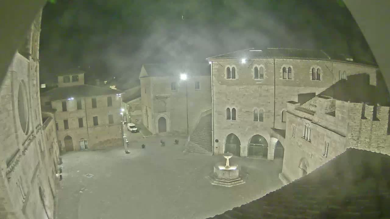 A nighttime view of a town square with several stone buildings surrounding a central fountain, under hazy or foggy conditions.