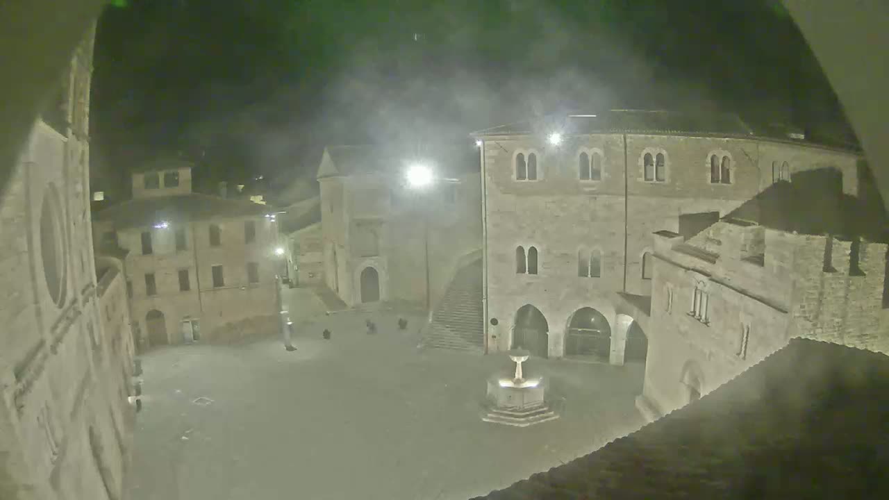 A nighttime, hazy view of a town square with several stone buildings surrounding a central fountain.