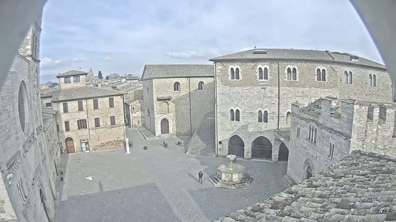 A stone plaza with a fountain is surrounded by medieval-style buildings under a partly cloudy sky.